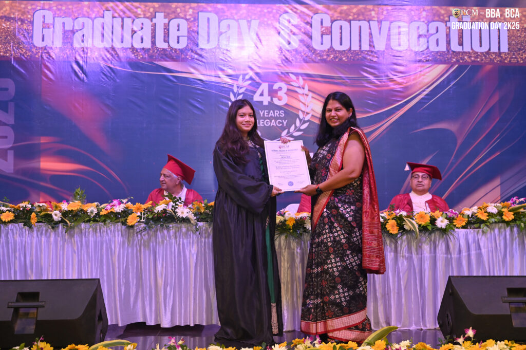 Two women on stage at a graduation ceremony, holding a certificate, banner in background.
