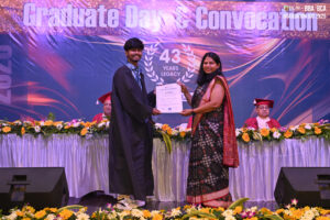 Graduate receives certificate at convocation; backdrop displays "43 Years Legacy," floral decor.