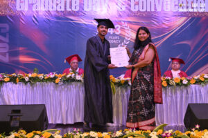 Graduate receiving a certificate from a woman in a saree on a decorated stage during the ceremony.