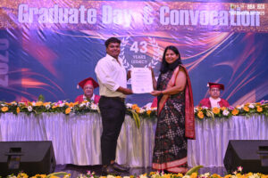 Graduate receives certificate on stage at convocation; decorated with flowers and banner.