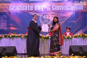 Man in gown and woman in saree exchanging a certificate on stage at graduation ceremony.
