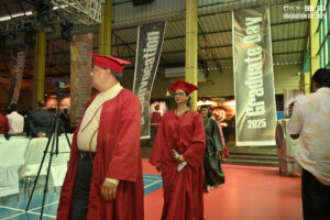 Individuals in red gowns at indoor graduation ceremony with "Graduate Day 2025" banners.