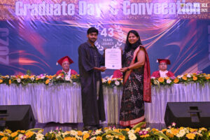 Man receives certificate from woman on flower-decorated stage; backdrop reads "Graduate Day & Convocation," "43 Years Legacy."