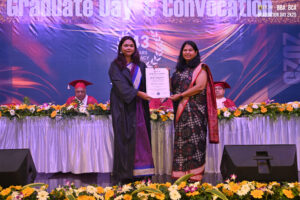 Graduate receiving certificate from woman in saree on stage, with floral decor and academic gowns.
