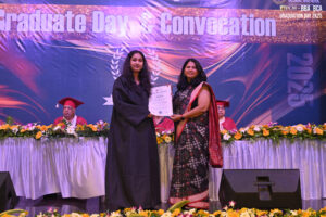Graduate in black gown receives certificate from woman in sari on stage during convocation.
