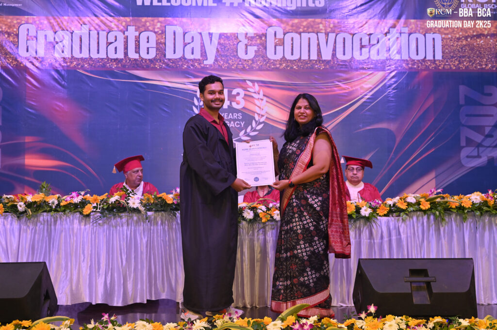 Man in graduation attire receives certificate from woman in saree at convocation ceremony stage.