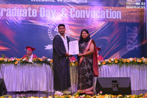 Graduate in gown receives certificate from woman in sari on stage; "Graduate Day & Convocation".
