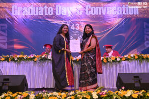 Graduation ceremony with two women holding a certificate, decorated table, and faculty on stage.