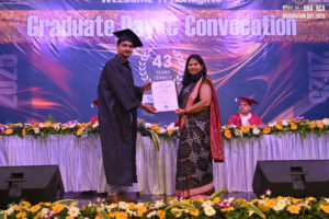 Graduate receives a certificate at convocation; banner and flowers decorate the stage.