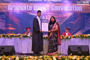 Graduate receives certificate from woman in traditional attire at ceremony, with faculty seated.