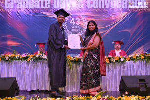 Man in graduation attire receives certificate from woman in traditional dress on decorated stage.