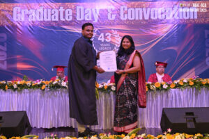 Graduate in cap and gown receives certificate, stage with "Graduate Day & Convocation" backdrop.