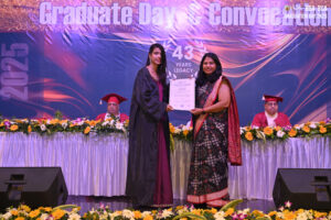Graduate in cap and gown receives certificate on stage, with decorated background and seated officials.