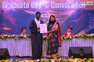 Graduation ceremony with graduate receiving certificate, decorated stage backdrop visible.
