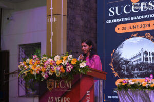 Woman speaking at graduation podium with flowers, display shows date June 28, 2024.