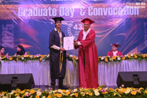 Graduation ceremony with graduate receiving certificate on decorated stage, banner above.