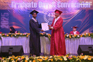 Graduate receiving diploma on stage at 2025 convocation, with decorative banner and flowers.