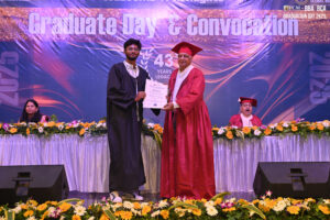 Graduate in black gown receives certificate on stage, with "Graduate Day & Convocation" backdrop.