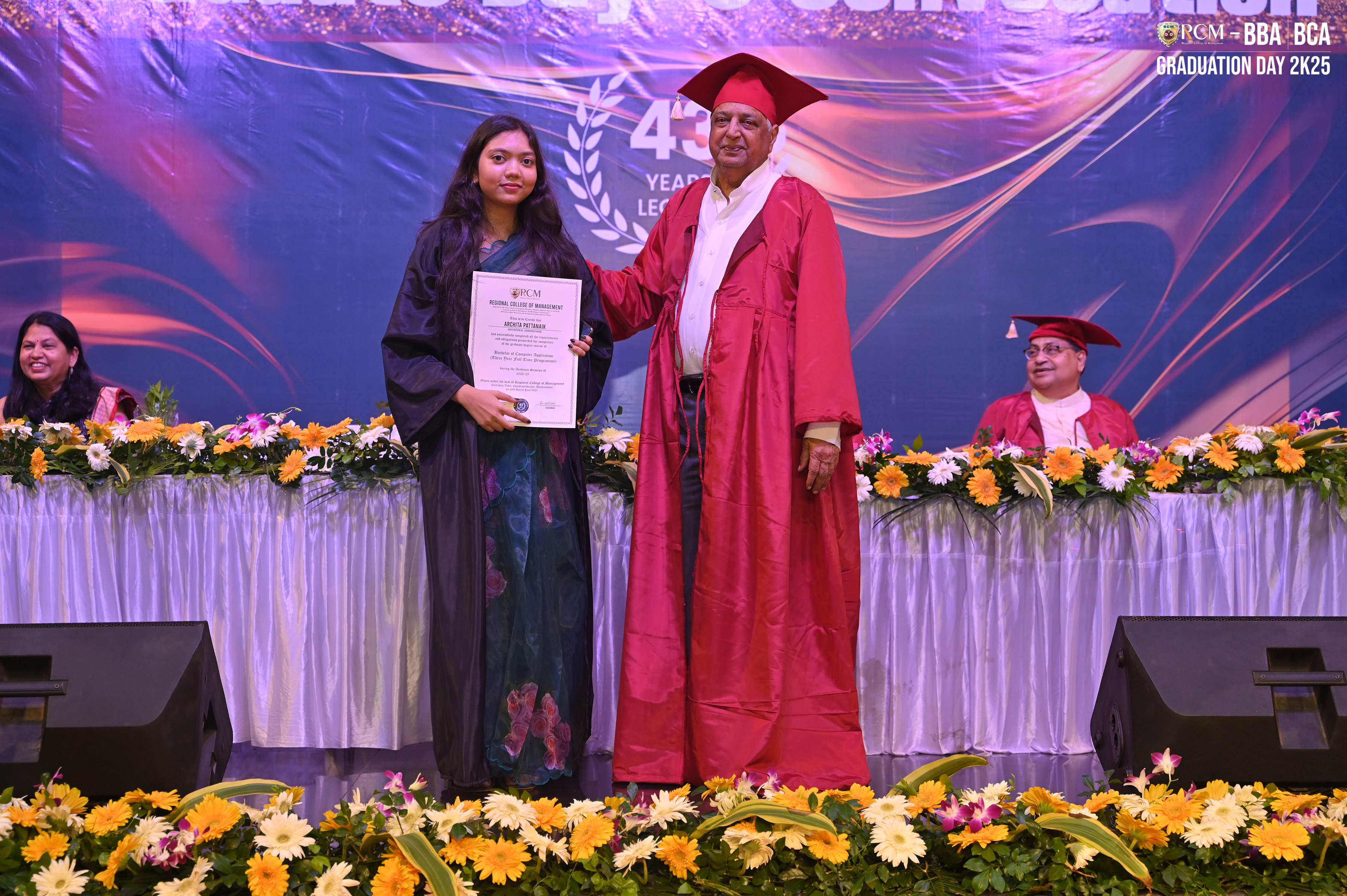 Graduate receives a certificate on stage with a decorated backdrop and seated individuals.