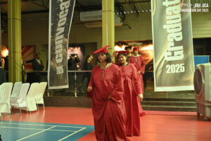 Graduates in red gowns walk in an indoor venue with "Graduation 2025" banners.