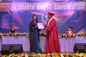 Graduation ceremony on stage; woman receives certificate from man in academic gown and cap.