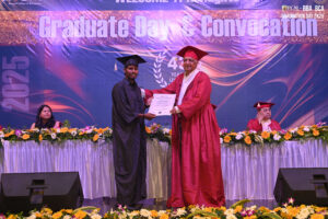 Graduate receives certificate from official on stage, backdrop reads "Graduate Day & Convocation 2025".
