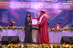 Graduate receiving certificate on stage; decorated with flowers; event banner visible.