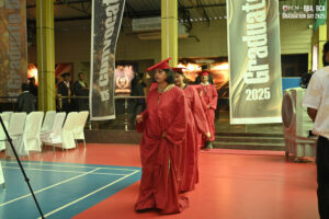 Individuals in red graduation gowns walking in line at an indoor graduation ceremony, 2025.