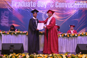 Graduate receiving certificate from faculty on stage at convocation, adorned with flowers.