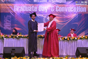 Graduate receives certificate from official on stage at convocation ceremony, flowers displayed.