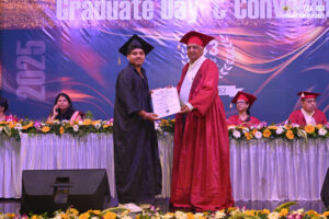 Graduate receiving certificate at 2025 ceremony, both in caps and gowns, banner and flowers visible.