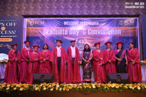 Graduates in red gowns and caps on stage, backdrop reads "Graduate Day & Convocation."