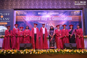 Individuals in red graduation attire on stage at RCM Graduate Day with banners and flowers.