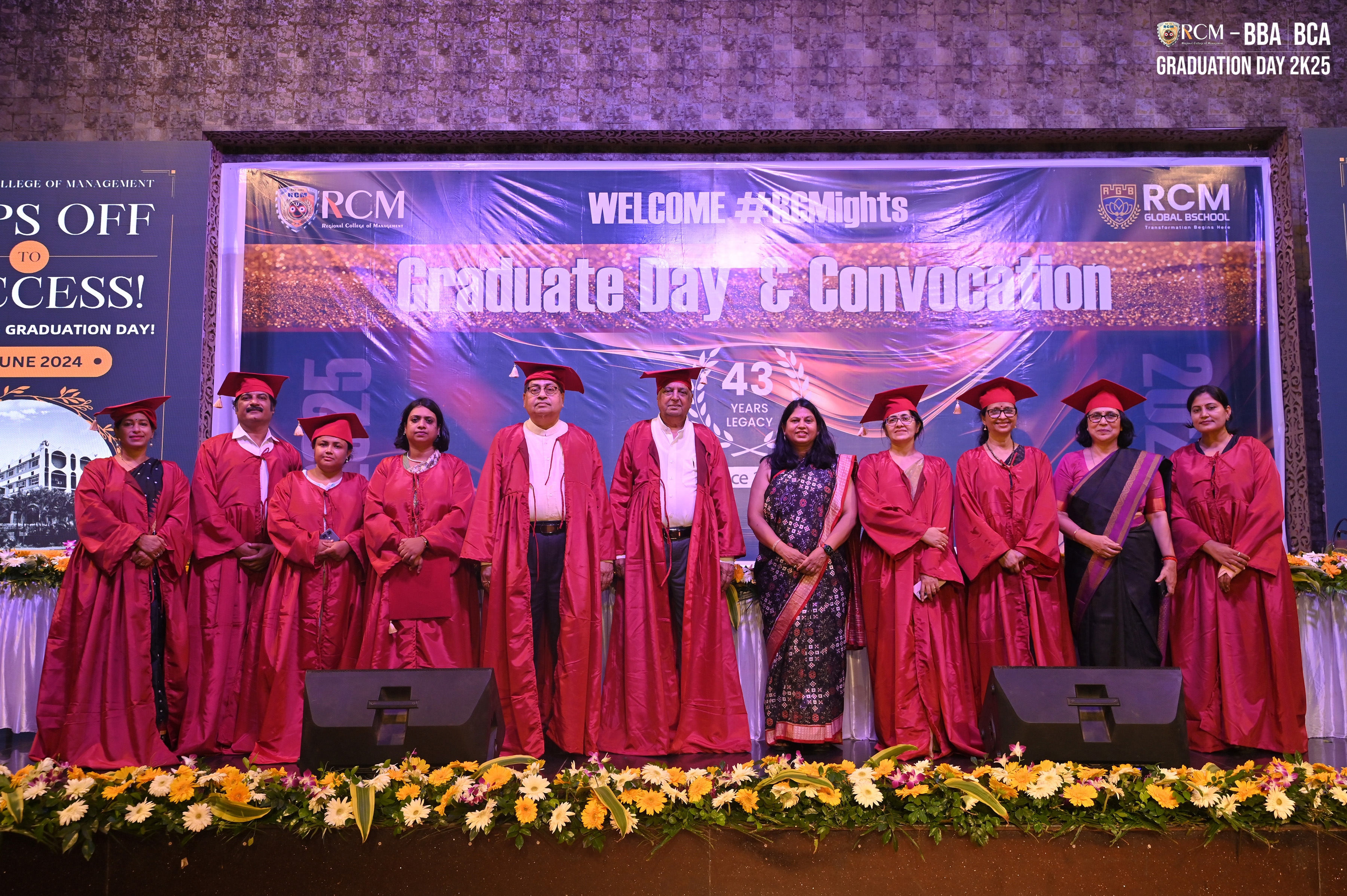 Individuals in red graduation attire on stage at RCM Graduate Day with banners and flowers.