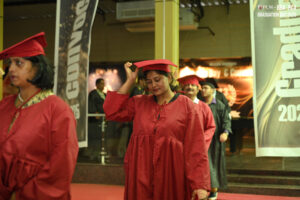 Graduates in red caps and gowns at a ceremony, woman adjusts cap, banners say "Graduation Day 2K23".