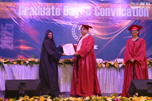 Graduate receiving certificate on stage at 2025 convocation with banner and flowers.