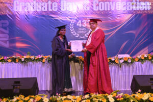 Graduate receives certificate on stage during ceremony with floral decorations and celebratory text.