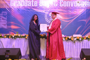 Woman in graduation gown receives certificate from man in red robe on decorated stage.