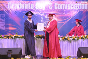 Graduate receiving certificate from faculty on stage during convocation, with flowers and banner.