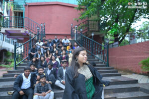Graduates on outdoor steps, one in traditional outfit; brick walls and greenery visible.