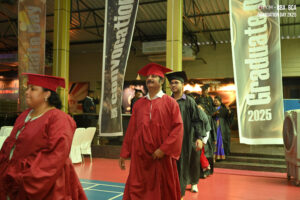 Graduates in red and black gowns walk indoors during 2025 ceremony with banners and decor.