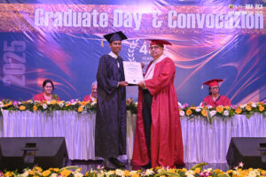 Graduate receives certificate from faculty during ceremony on decorated stage with banner.