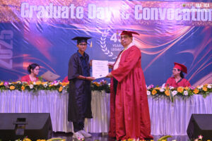 Graduate receives certificate on stage during convocation; two seated in red gowns at flowered table.
