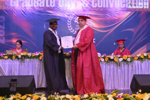 Graduate in cap and gown receives certificate from person in red robe on decorated stage.