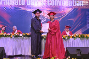 Graduate in black receives certificate from faculty in red on stage; decorated table behind.