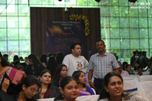 People seated indoors at a graduation event, with others standing and conversing nearby.