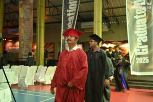 Graduates in caps and gowns at indoor ceremony with 'Graduate 2025' banners and empty chairs.