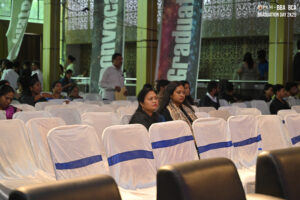 Graduation event with attendees seated on white chairs with blue sashes in a decorated hall.