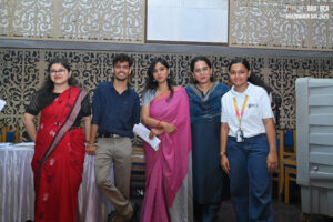 Five individuals pose indoors in formal attire, including saris and a polo, with ornate panels.