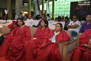 People in red gowns seated indoors at a graduation event in a large, well-lit hall.